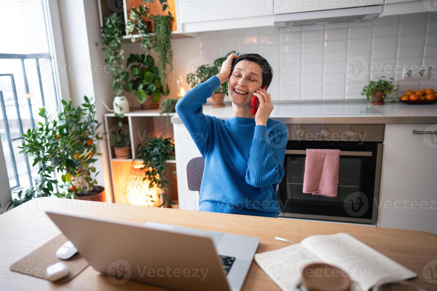 Smiling mature woman chats on phone, work on laptop and setting up online training sessions at home photo