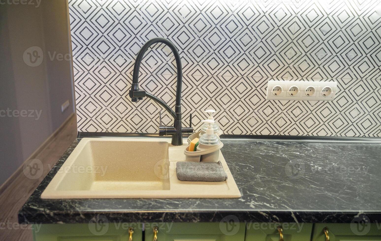 Close-up of a light beige kitchen sink with a modern black faucet, dark green marble countertop, and a black and white tile backsplash. Dishwashing bottles are nearby. photo
