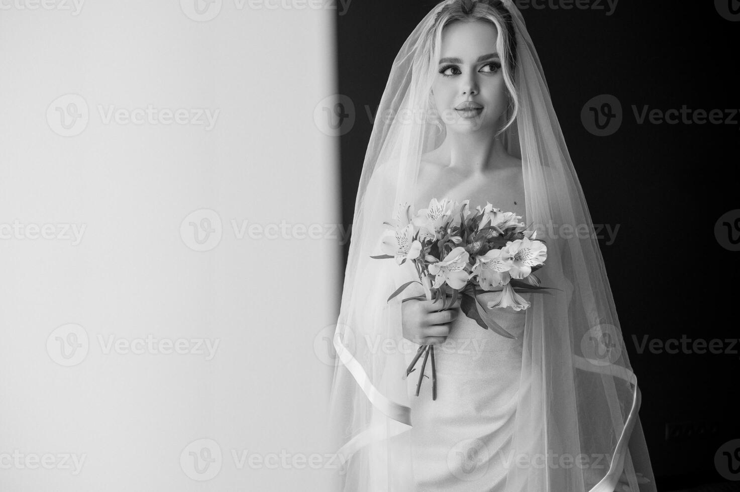 portrait of a blonde bride with a lush veil, in a long white dress near the window with a bouquet of lilies. Minimalistic interior, hotel or house. bride in a long dress and high hairstyle. black and white photo