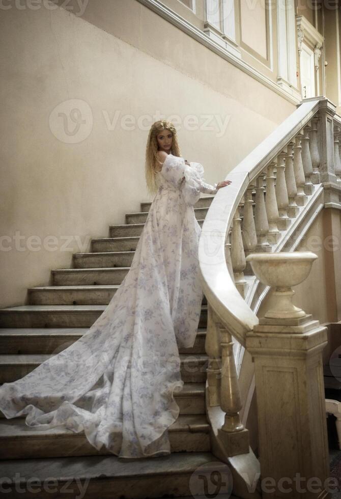 beautiful blonde with long hair in long light fantasy ball gown like fairy princess. She is walking in stone palace castle with balconies and stairs. photo