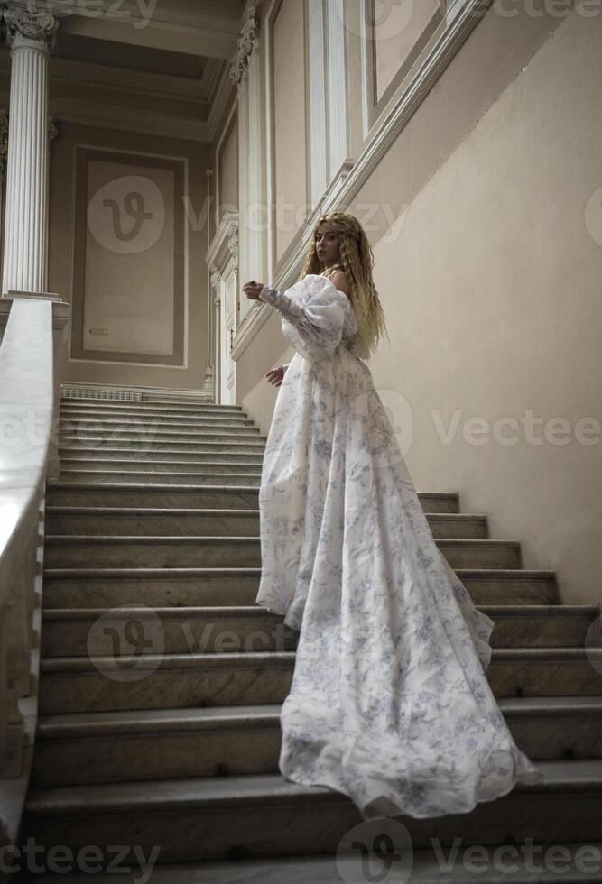 beautiful blonde with long hair in long light fantasy ball gown like fairy princess. She is walking in stone palace castle with balconies. standing on stairs going up stairs of romantic castle photo