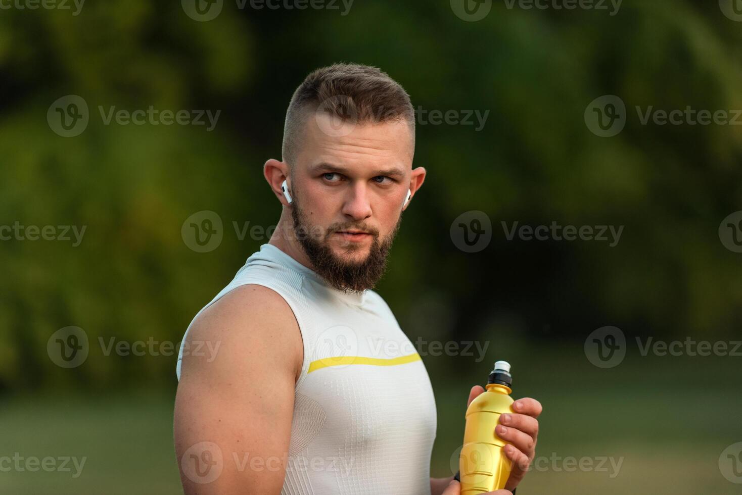 A young fit man listening to music on wireless headphones and drinking water or nutrition juice from a bottle after a run or workout in a summer park, active healthy lifestyle. photo