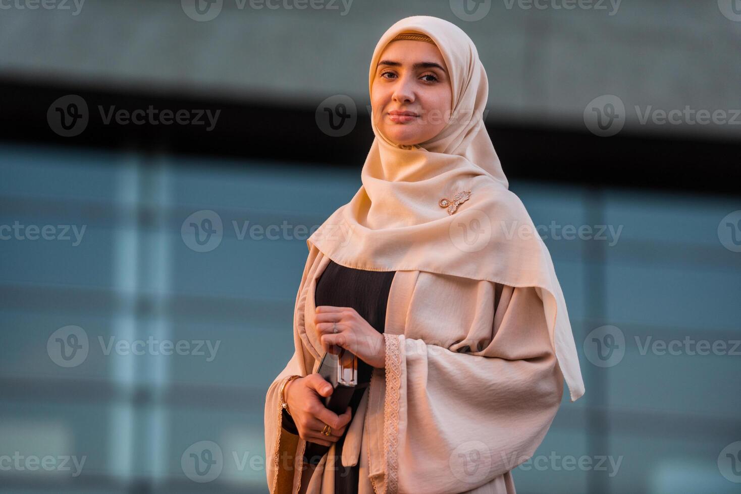 Portrait of a young confident Muslim business woman in hijab near the modern office building, looking at the camera with a friendly smile. photo