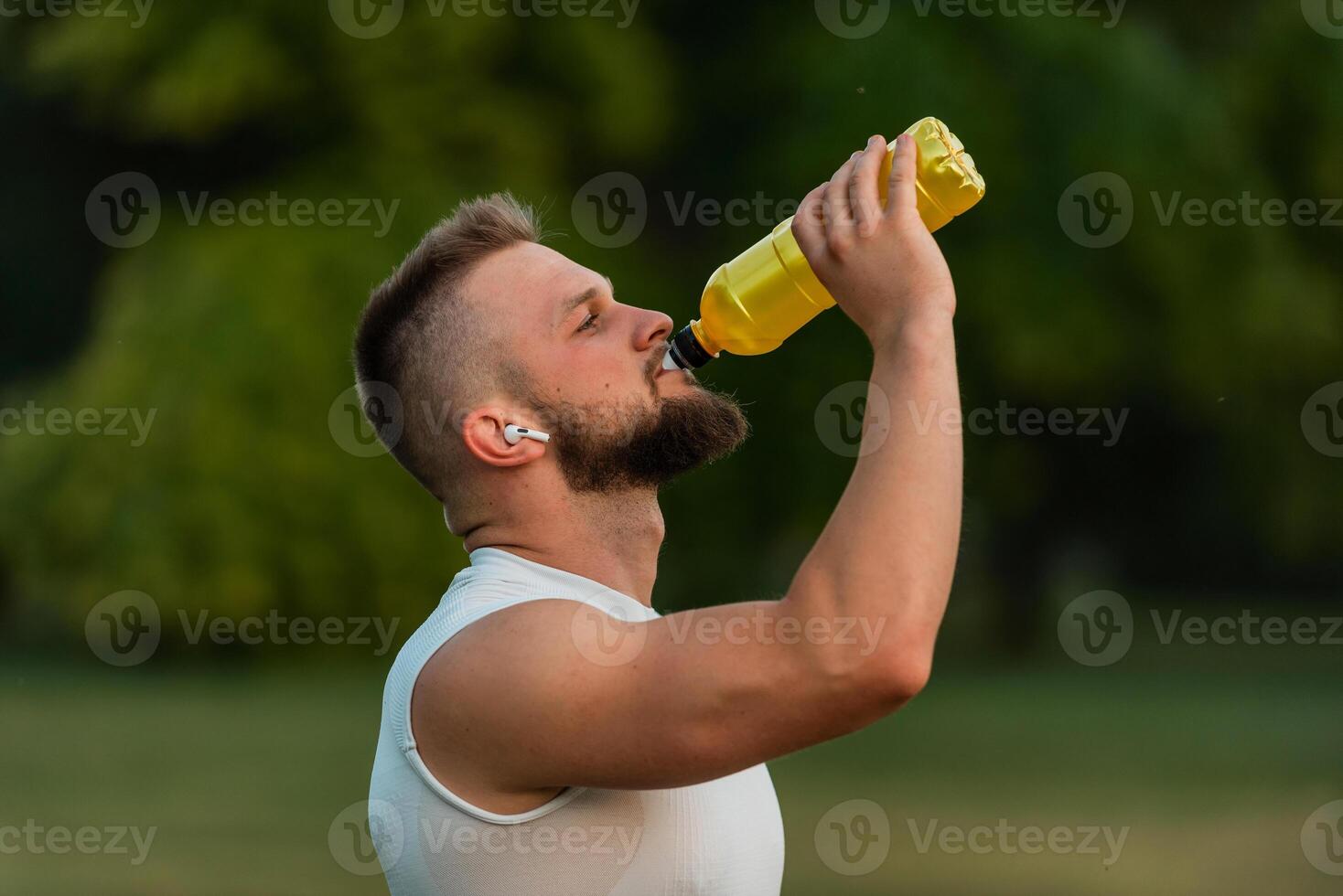 A fit young man with closed eyes listening to music on wireless headphones and drinking water or nutrition juice from a bottle after a run or workout in a park active healthy lifestyle. photo