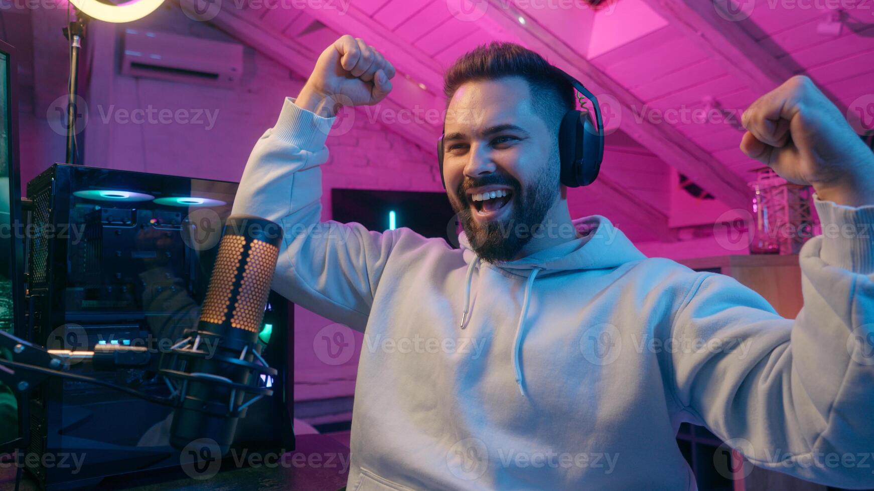 Happy young man Caucasian guy male celebrating victory gaming on computer stream with headphones and microphone in neon room joyful gamer excited after winning online battle rejoice reaction winner photo