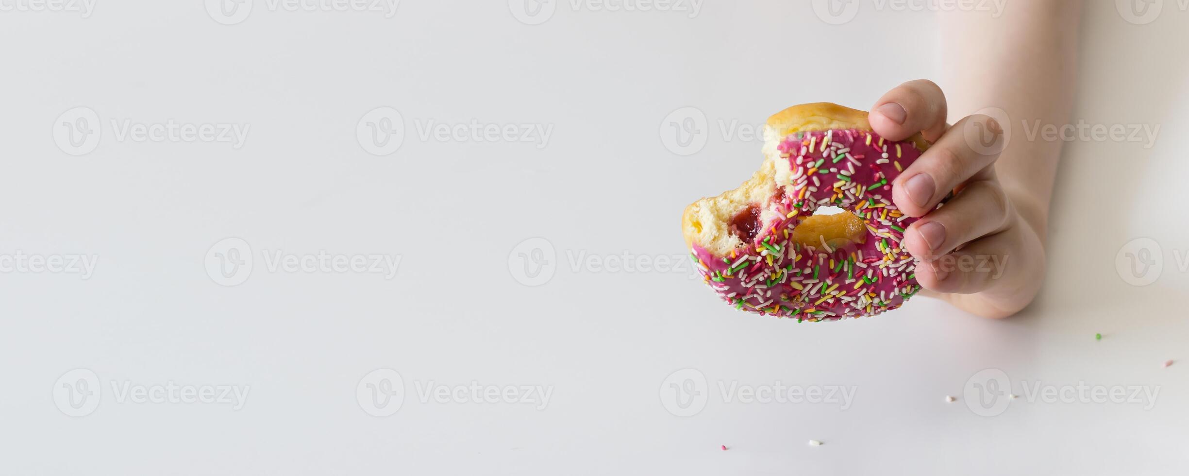 Children's hand stretches a bitten donut in a pink glaze. Banner format photo