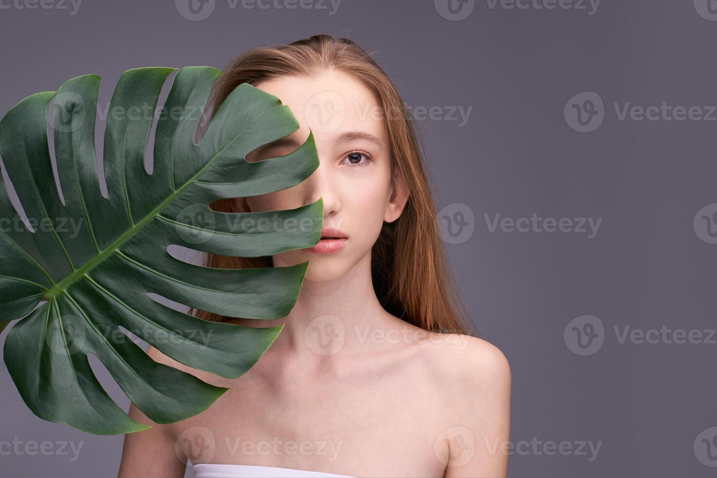Young caucasian female with long hair holding large green leaf against gray background. photo