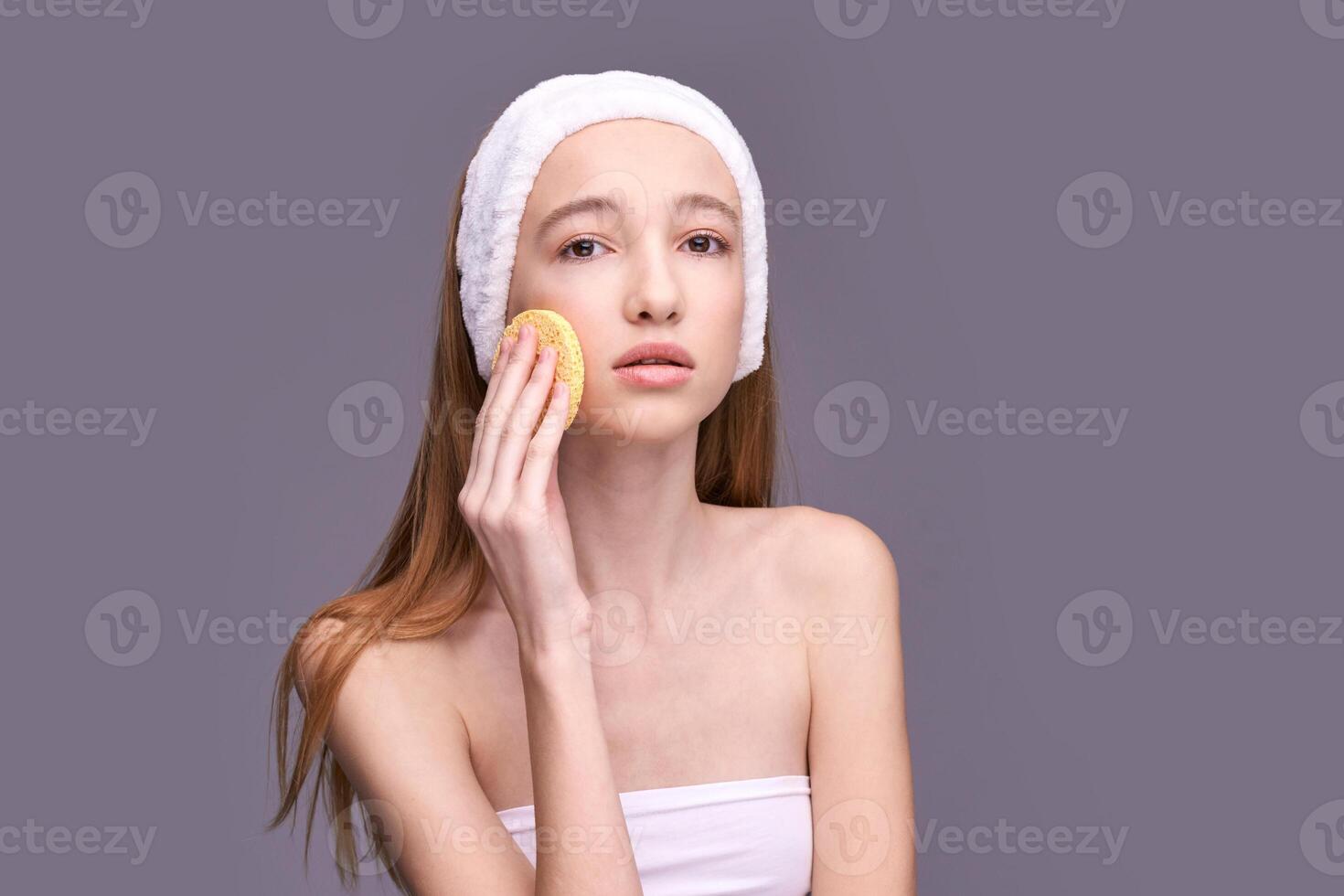 Young caucasian female with spa headband cleansing skin using facial sponge against gray background. photo