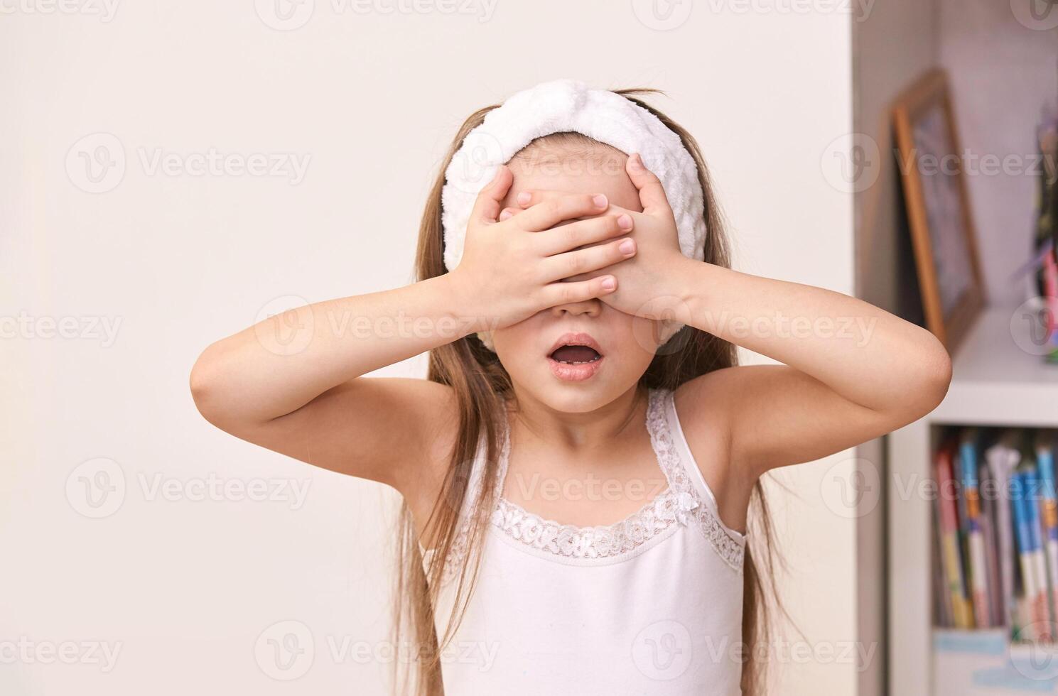Caucasian female child in white tank top covering eyes with hands in playful pose. photo