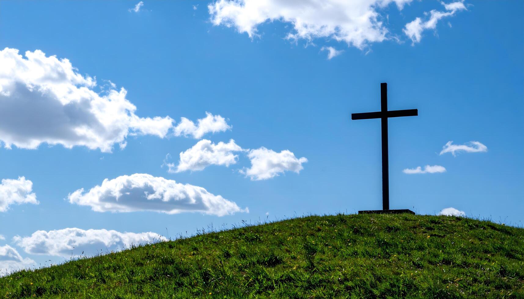 Silhouette of a cross standing on a grassy hill under a bright blue sky with white clouds. Perfect for ascension day jesus christ. photo