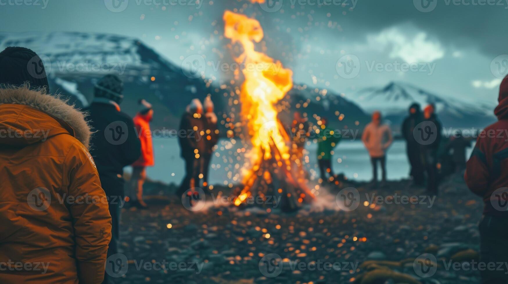 A group of diverse people celebrating Iceland Independence Day around a large bonfire. Snow-capped mountains and a lake are visible in the background. The atmosphere is festive and lively photo