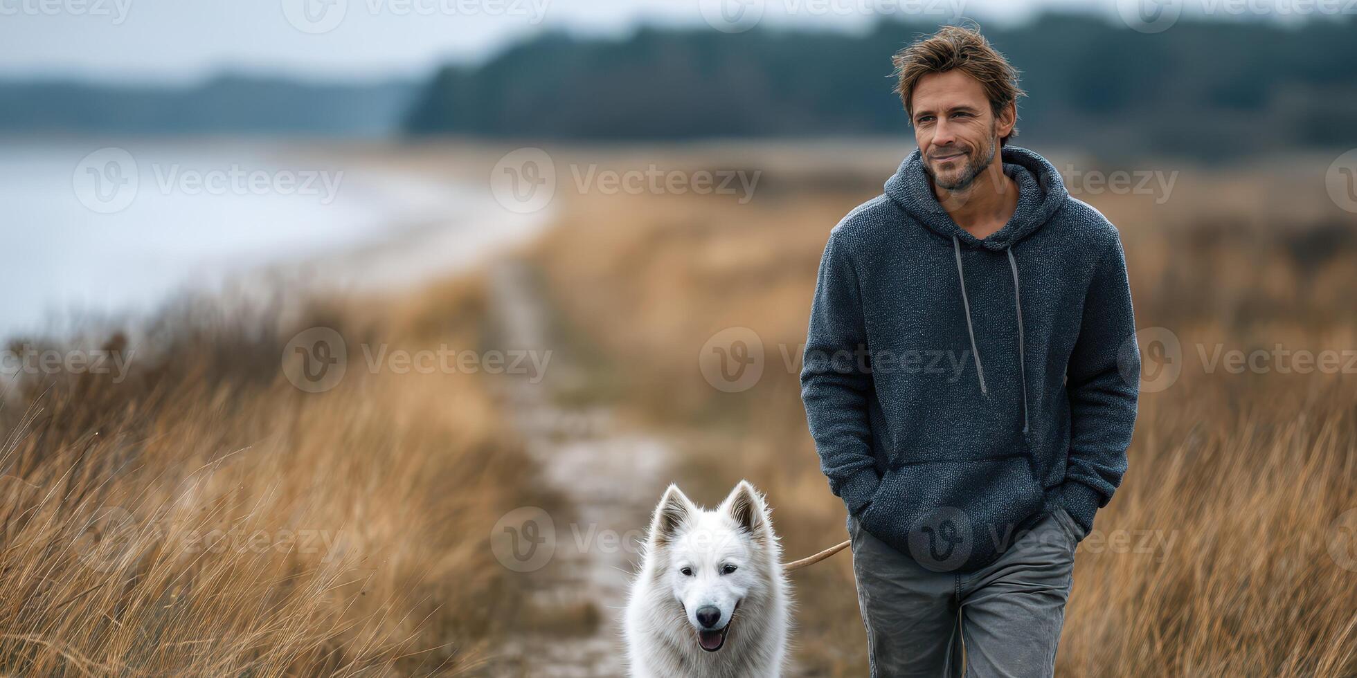 Man walking with a white dog along a grassy path near a serene lake during a cloudy afternoon photo