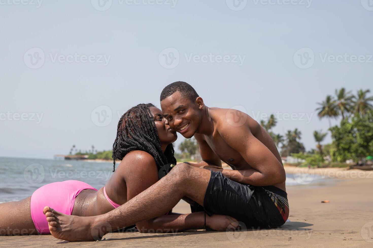 Young african couple kissing, smiling while sitting on the sand at the beach photo