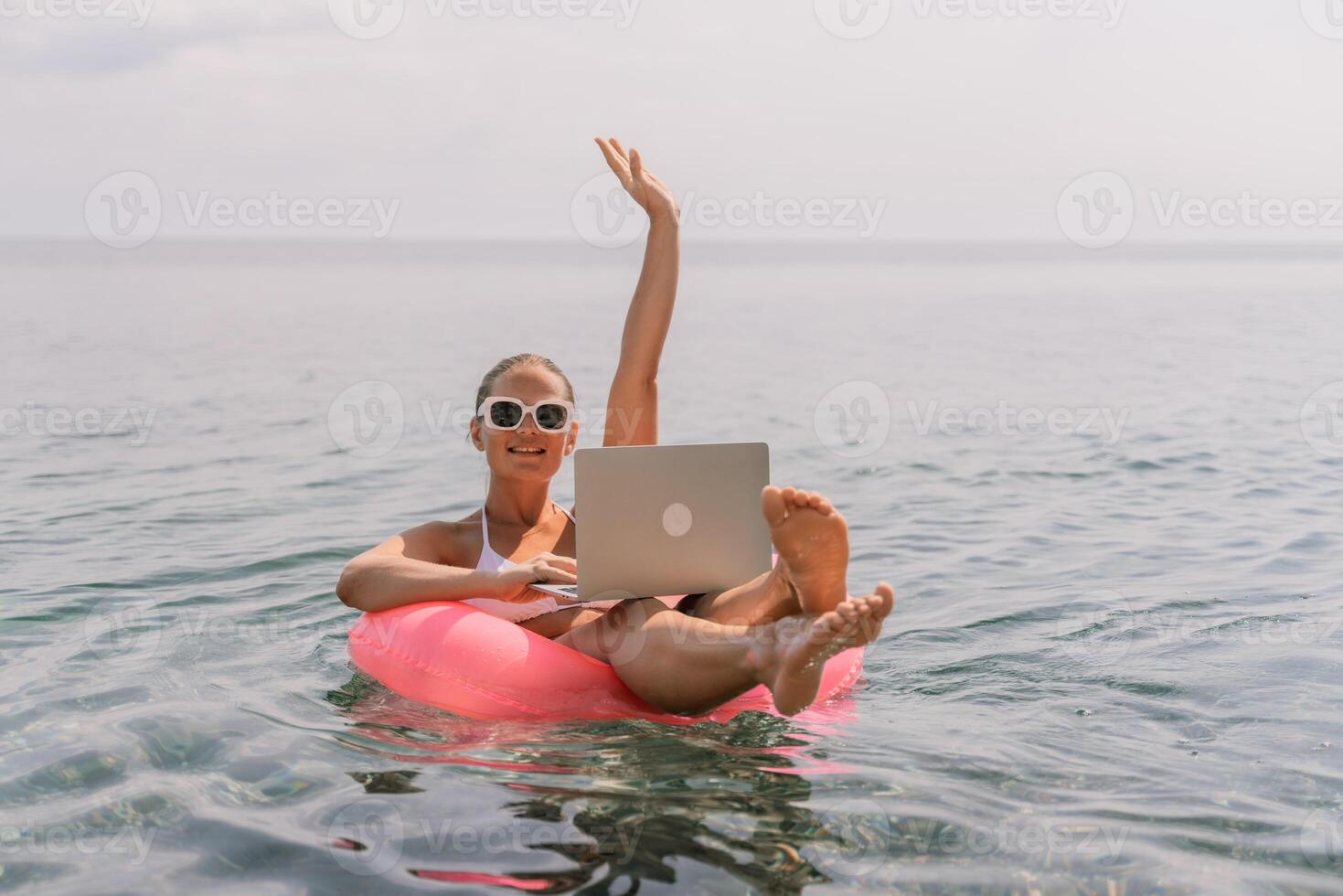 A woman is sitting on a pink inflatable raft in the ocean with a laptop on her lap. She is smiling and she is enjoying her time. photo