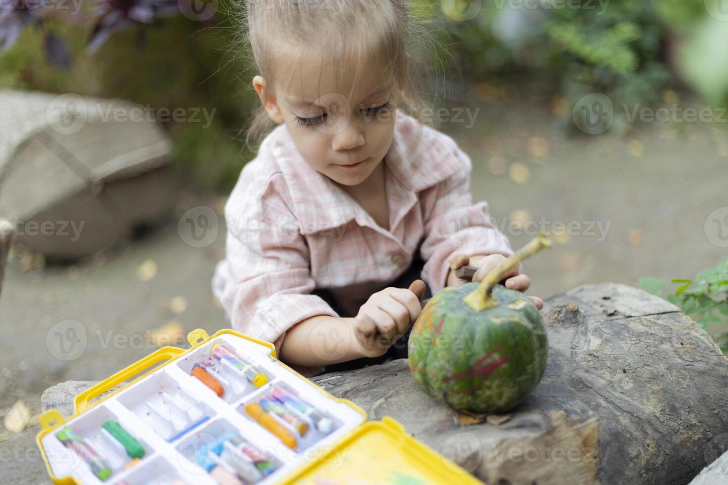 A little girl draws with concentration on a pumpkin while sitting in a green garden. Halloween preparation. Diy activities for children photo