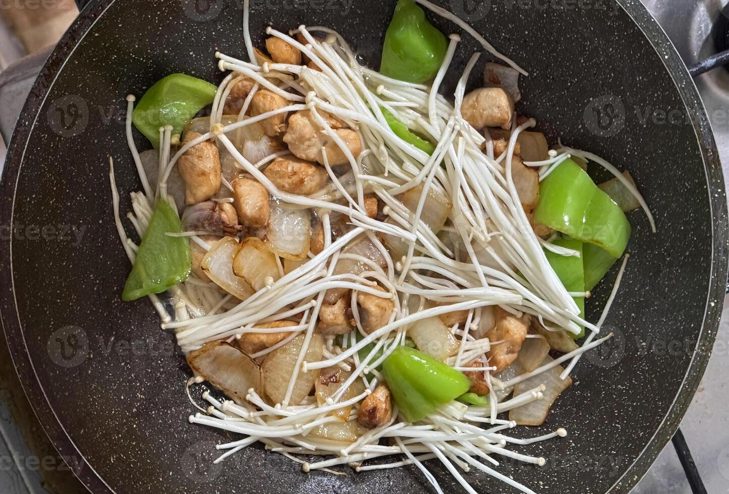A frying pan filled with enoki mushrooms, chicken pieces, bell peppers, and onions. A colorful and appetizing dish in the process of cooking photo