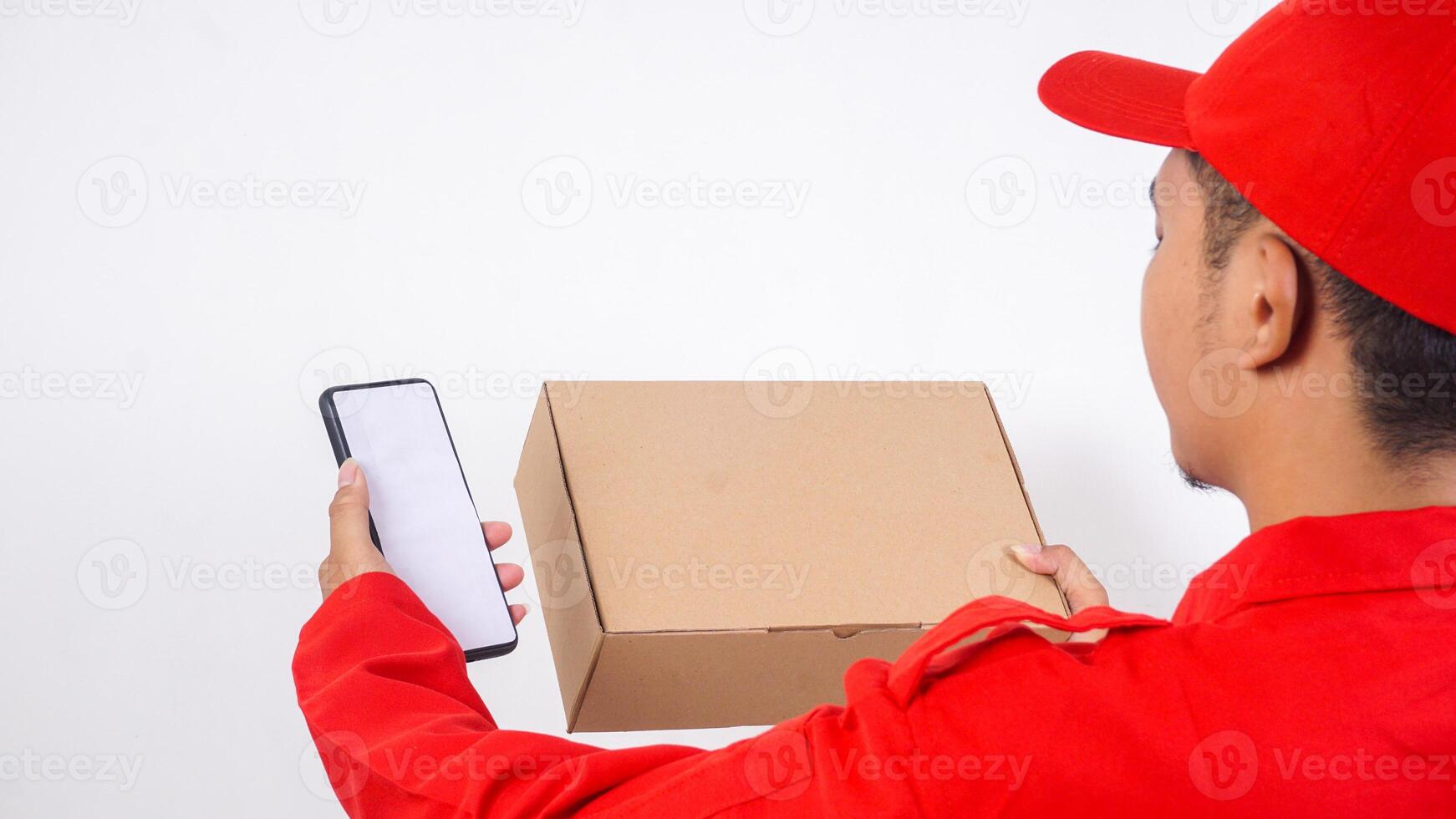Rear view of delivery man in red uniform holding a package box parcel and smartphone isolated on white background photo