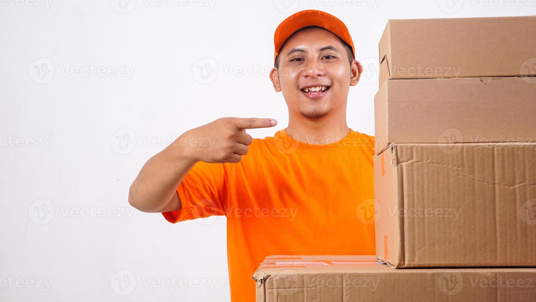 Happy Asian deliveryman in orange t-shirt uniform with stack of package boxes parcel and pointing at the boxes isolated on white background photo