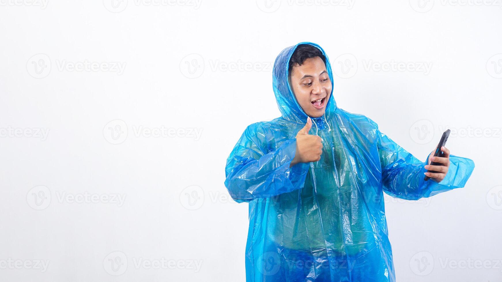 Smiling Asian young man in blue raincoat looking at smartphone and raising thumb up isolated on white background photo