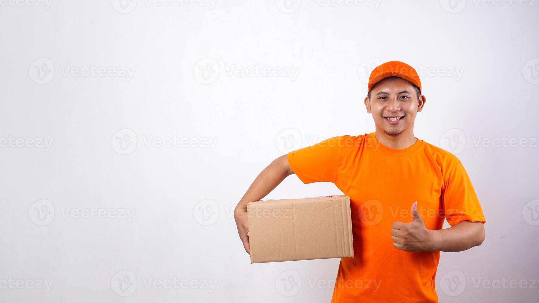 Asian deliveryman in orange t-shirt uniform carrying a package box parcel and showing his thumb upisolated on white background photo