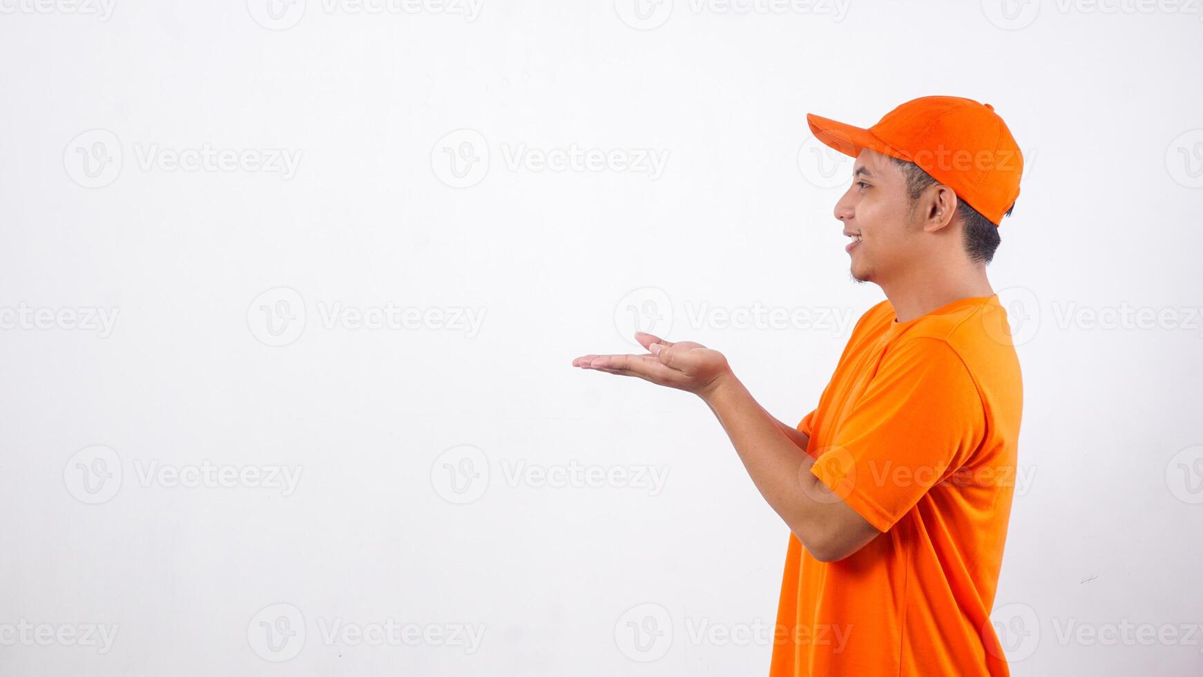 Side view of Asian deliveryman in orange t-shirt uniform poses as if he is presenting something in empty space area isolated on white background photo