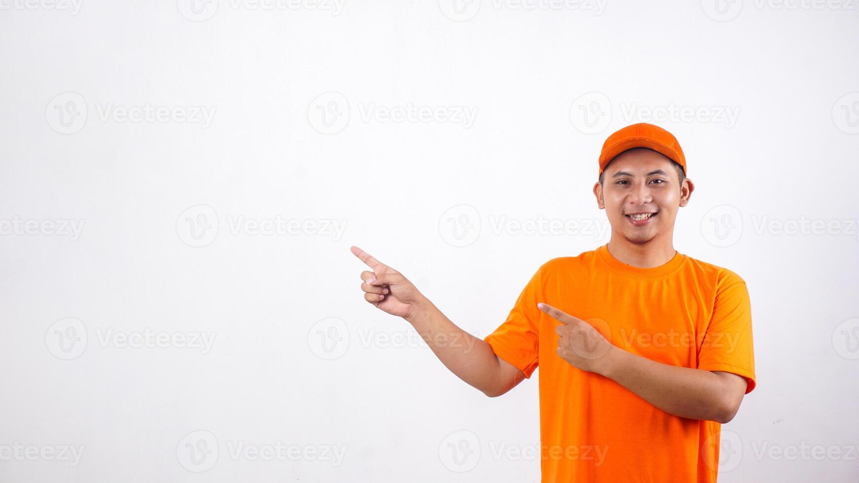 Smiling Asian deliveryman in orange t-shirt uniform poses as if he is pointing something in empty space area isolated on white background photo