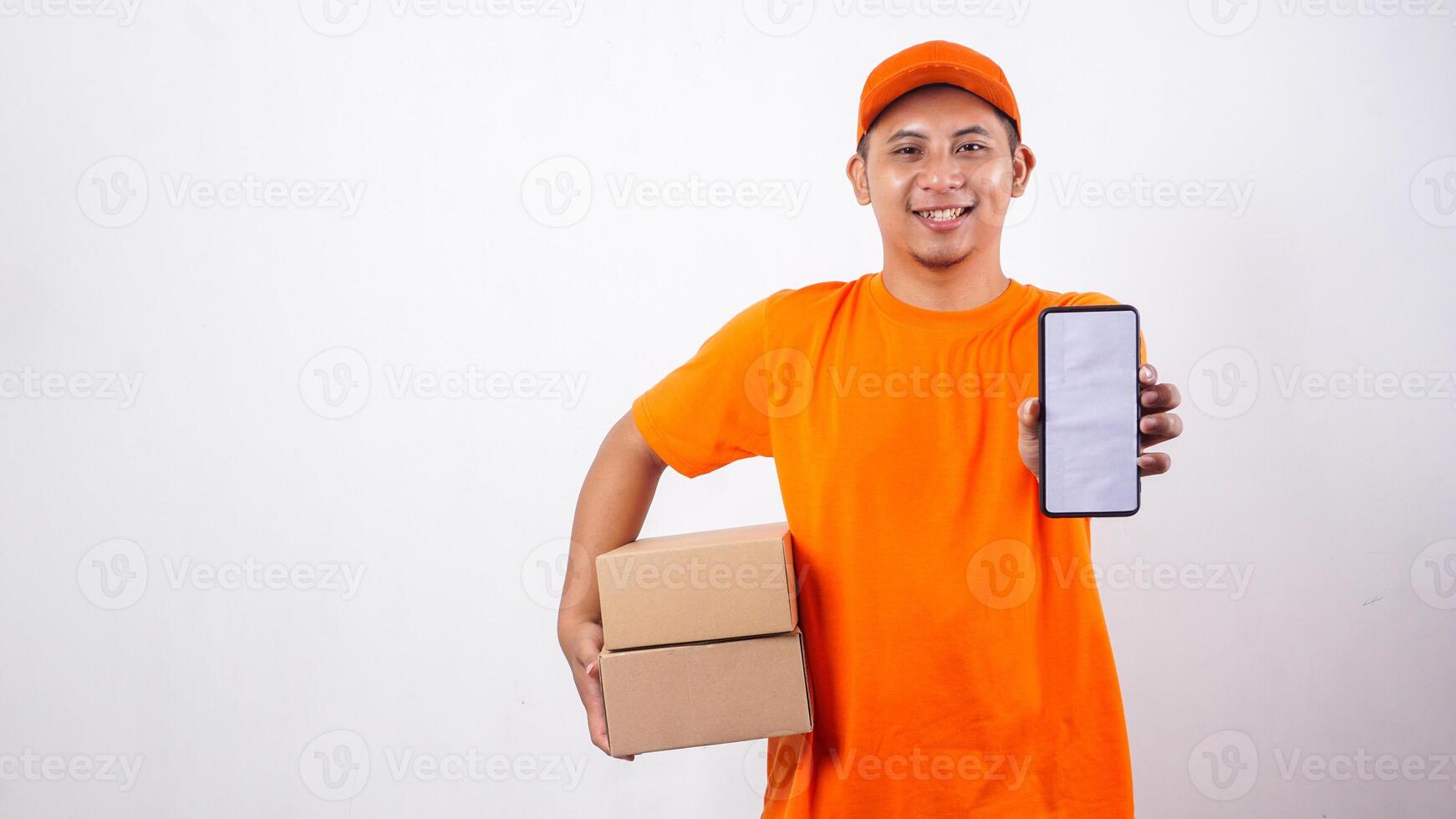 Portrait of Asian Delivery man in orange t-shirt carrying package boxes parcel while showing a smartphone with blank screen isolated on white background photo