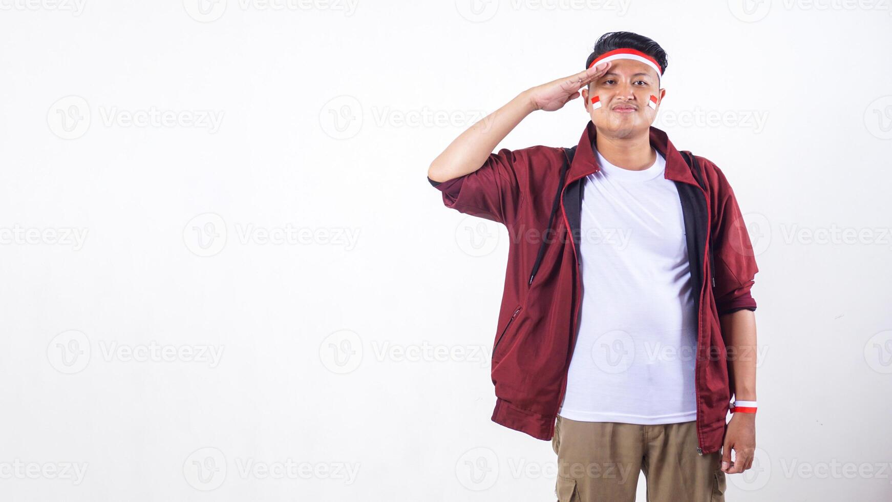 Smiling Indonesian man with red and white ribbon wristband doing salute pose to celebrate independence day of Indonesia isolated on white background with copy space photo