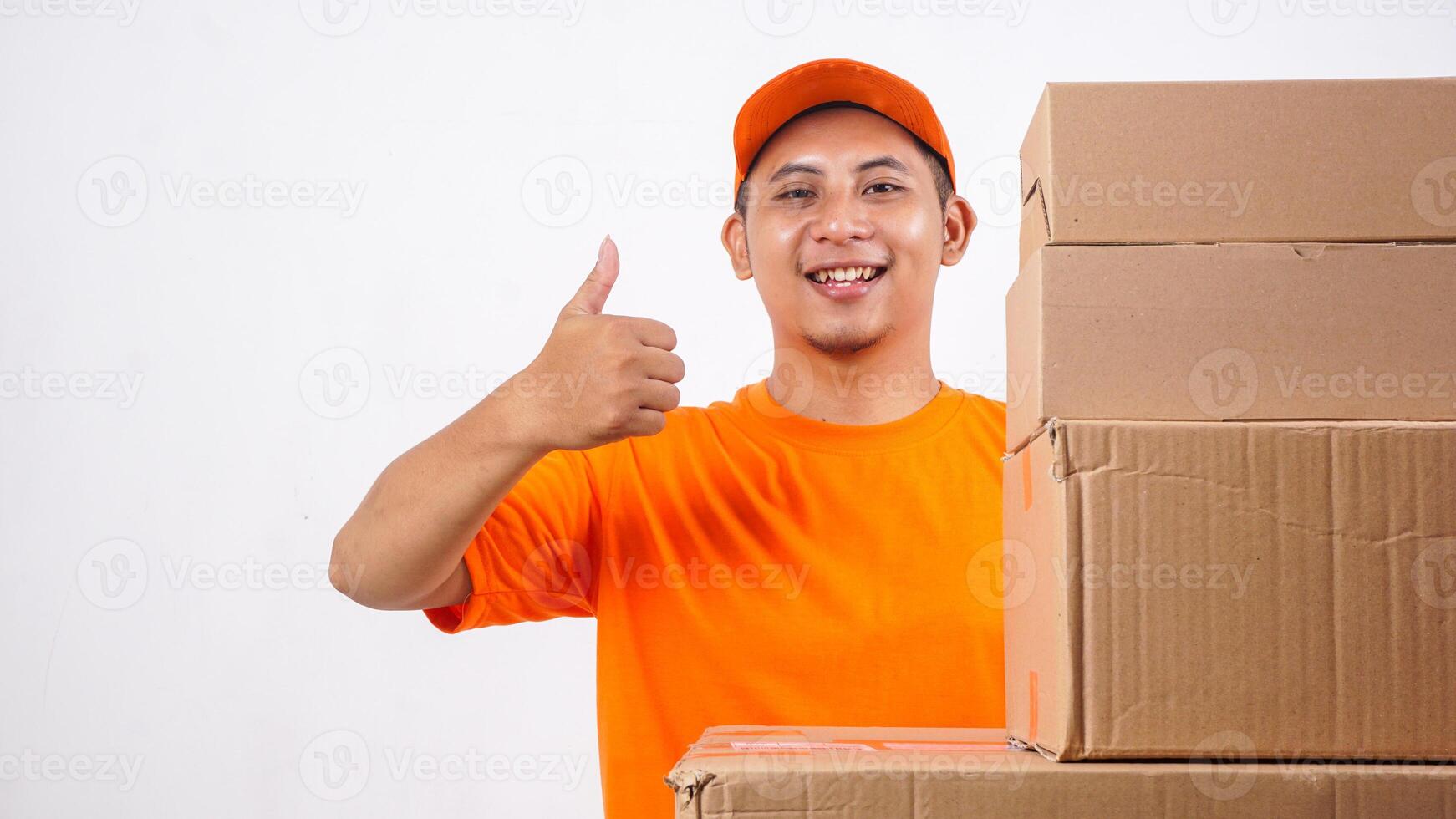 Happy Asian deliveryman in orange t-shirt uniform with stack of package boxes parcel and showing his thumb up isolated on white background photo