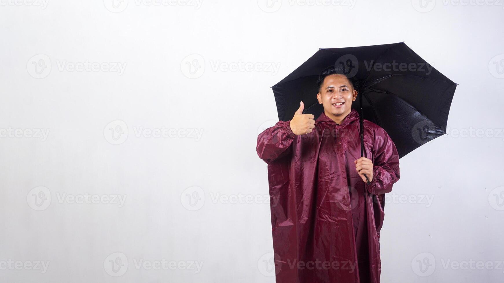 Smiling Asian man in red raincoat holds umbrella and showing his thumb up isolated on white background photo