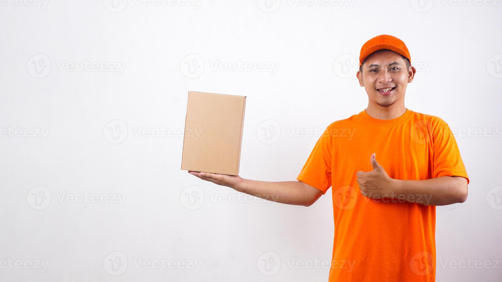 Asian deliveryman in orange t-shirt uniform with a package box parcel and showing his thumb up isolated on white background photo
