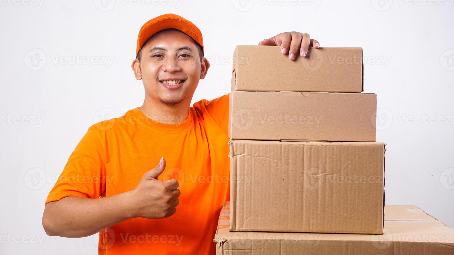 Smiling Asian deliveryman in orange t-shirt uniform with stack of package boxes parcel and showing his thumb up isolated on white background photo