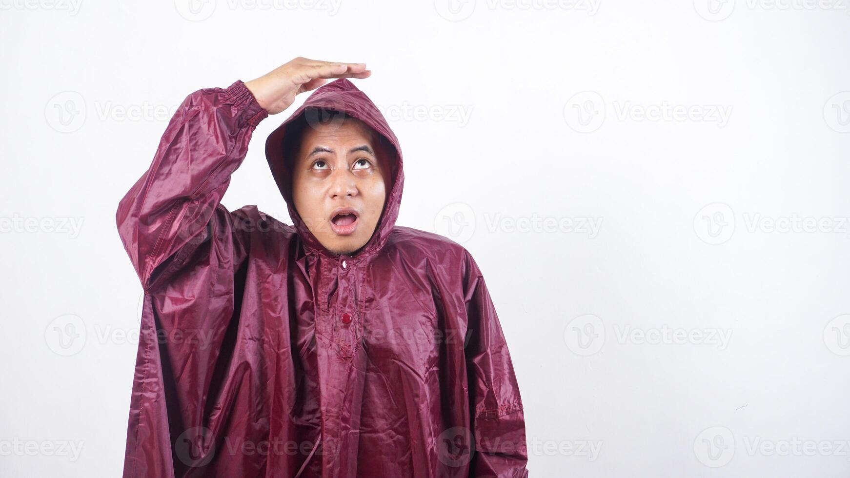 Portrait of Asian man in red raincoat covering his head with hands to shield from rain while looking up to the sky isolated on white background photo