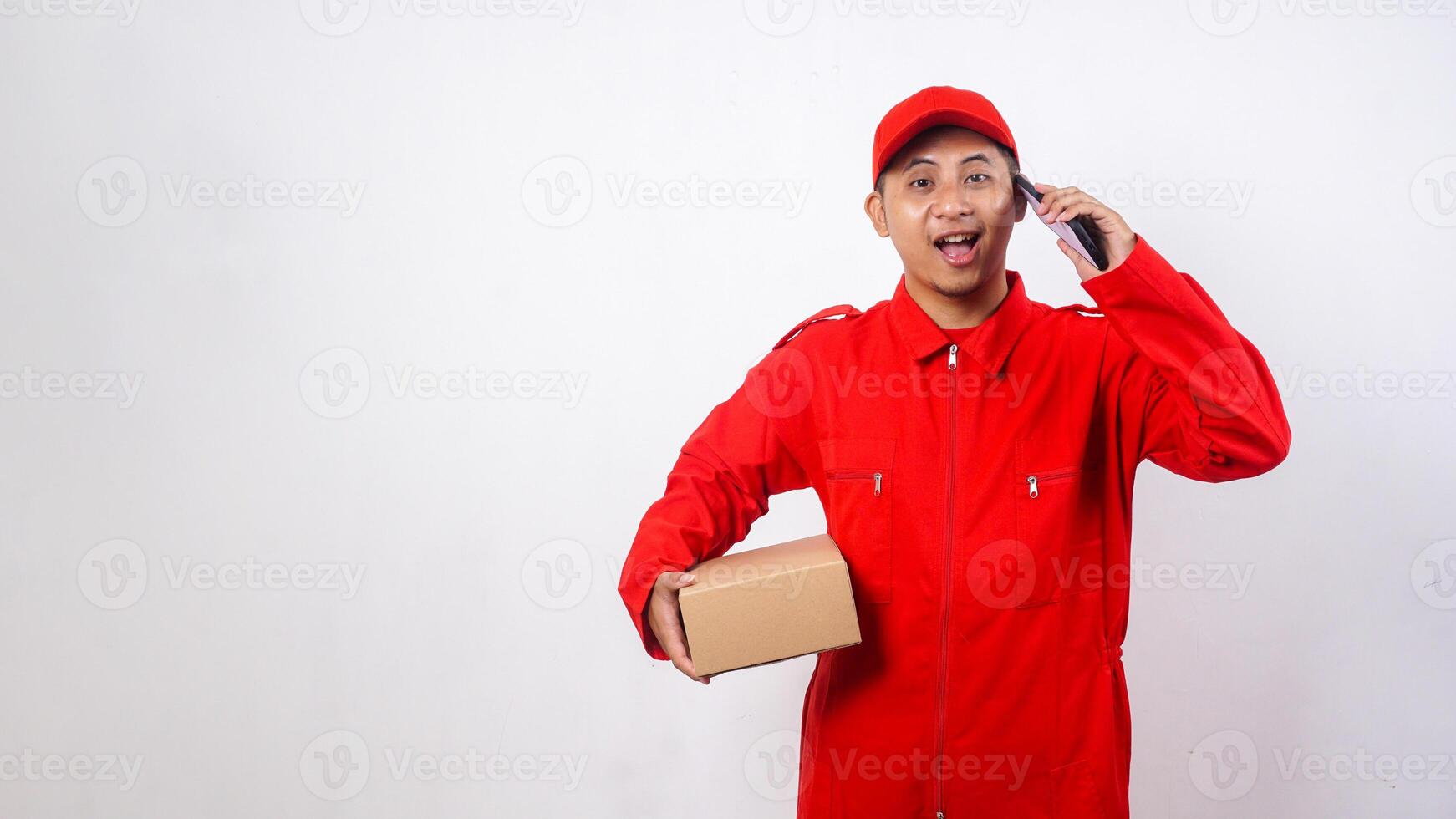 Asian Delivery man in red uniform carrying a package box parcel while taking a phone call isolated on white background photo