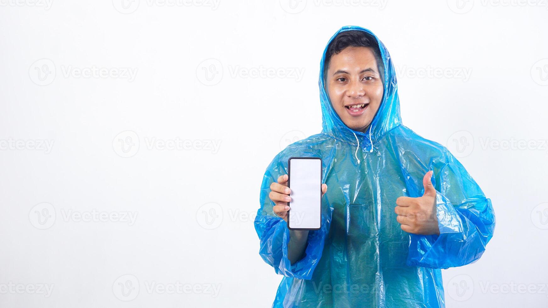 Portrait of Asian young man in blue raincoat showing smartphone mock up with blank screen and raising thumb up isolated on white background photo