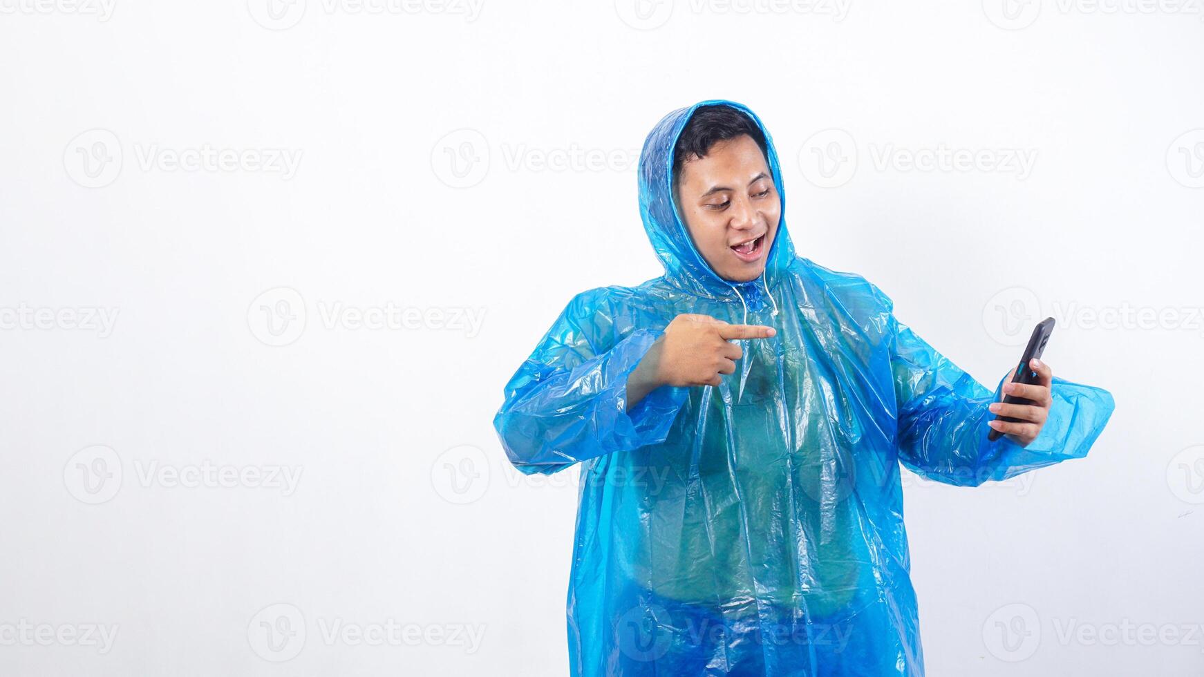 Smiling Asian young man in blue raincoat pointing at smartphone screen isolated on white background photo