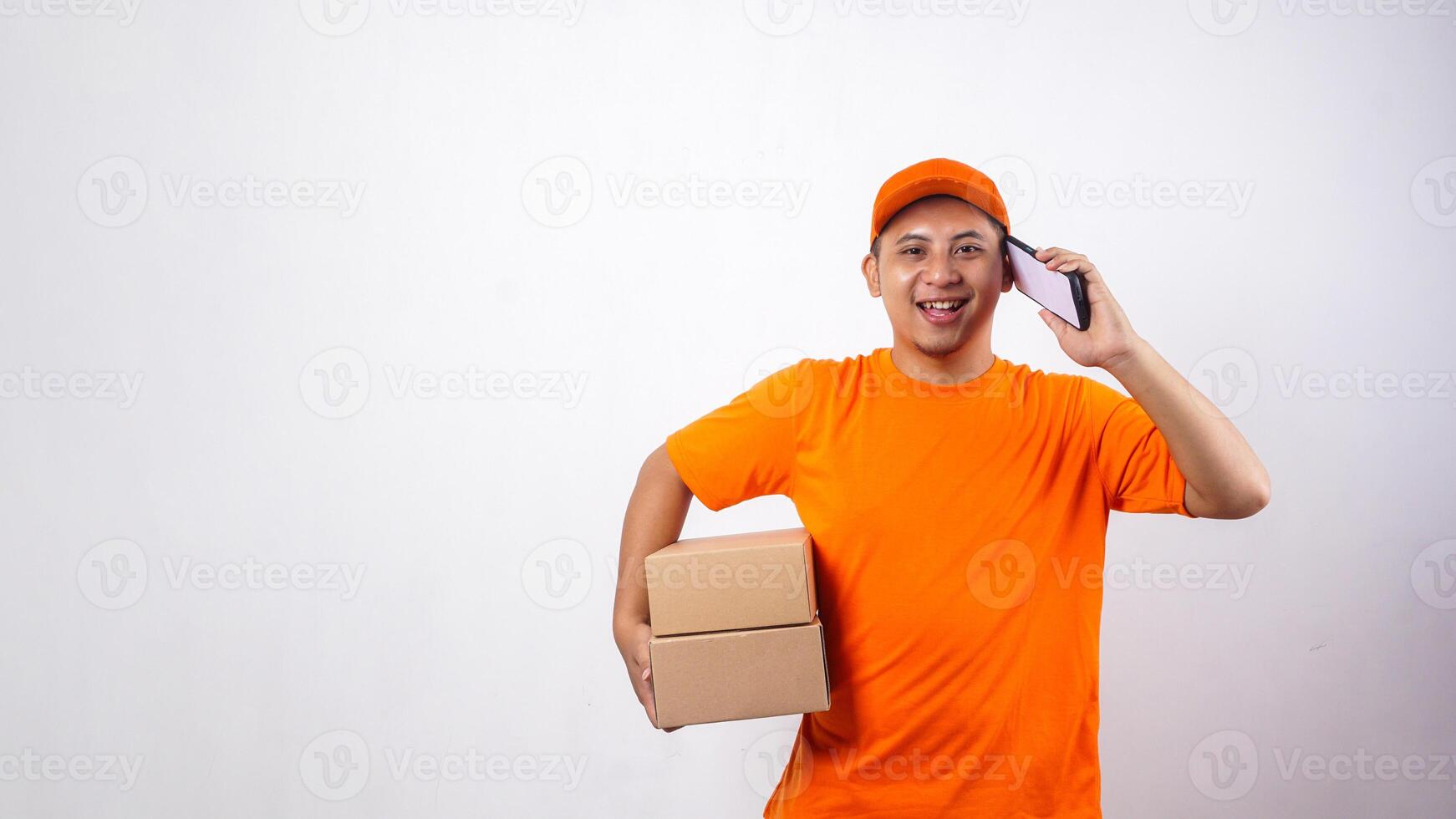 Asian Delivery man in orange t-shirt carrying package boxes parcel while taking a phone call isolated on white background photo