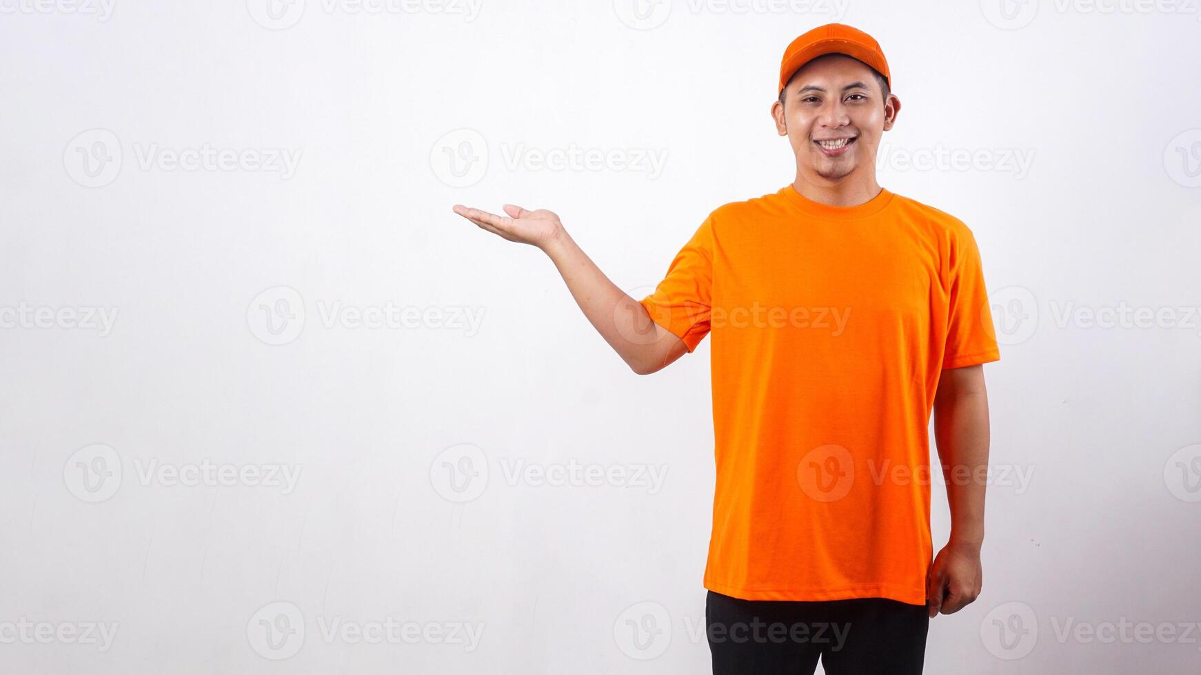 Smiling Asian deliveryman in orange t-shirt uniform poses as if he is presenting something in copy space area isolated on white background photo