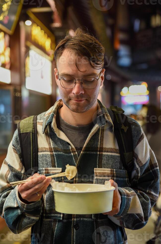 Russian tourist holding a crab and pork dumpling with chopsticks, looking at it before eating, on Yuyuan Old Street in Shanghai, wearing a plaid shirt photo