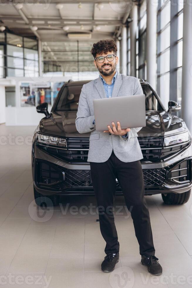 Car salesman holding laptop standing in front of new vehicle in car showroom dealership photo