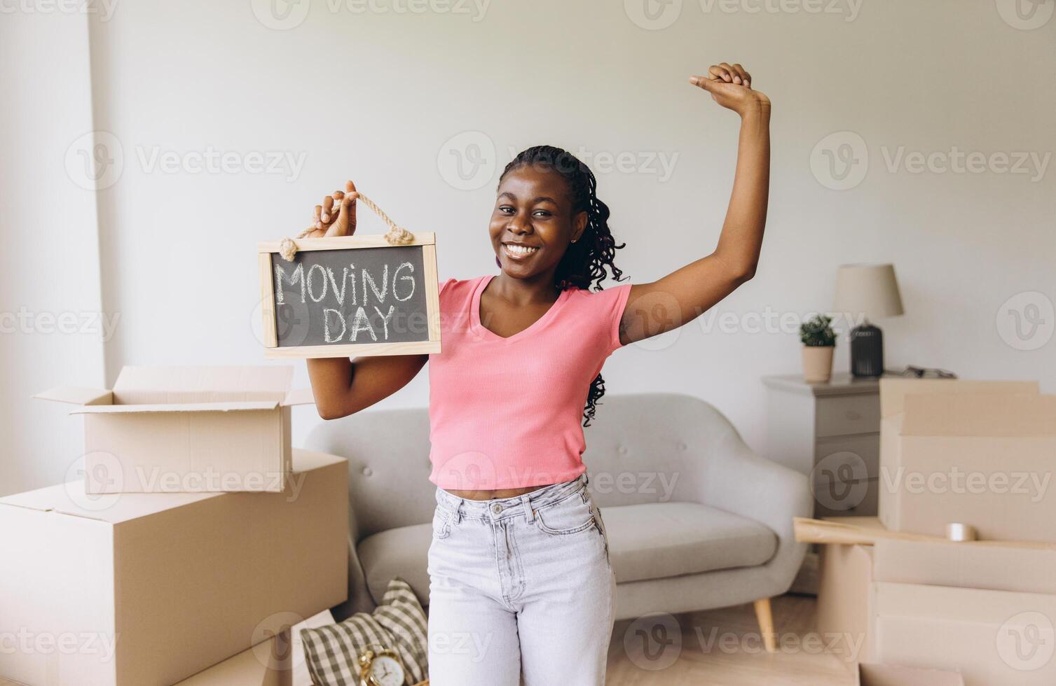 Happy African American woman holding moving day sign and dancing in new apartment photo