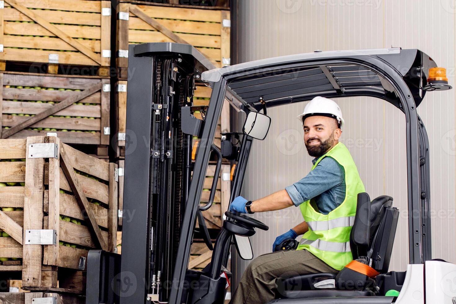 Employee on electric forklift carry container with ripe apples at storage. Smiling millennial bearded man drives in hard hat in truck, lifts up fruit, on many wooden crates at warehouse, free space photo