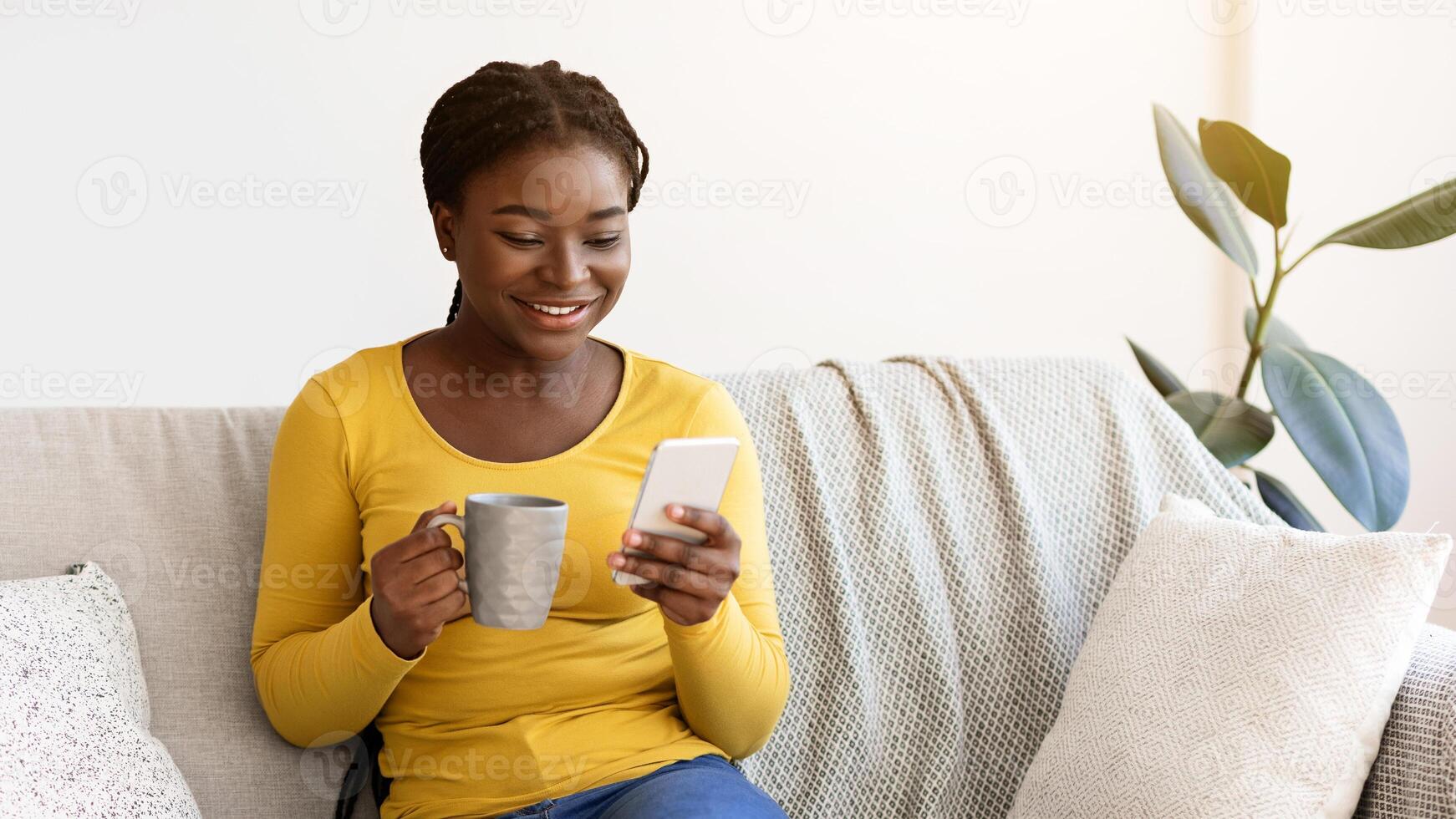 Domestic Relax. Positive black lady resting on couch with smartphone and coffee, browsing internet and enjoying hot drink, african woman checking social networks or messaging with friends, copy space photo