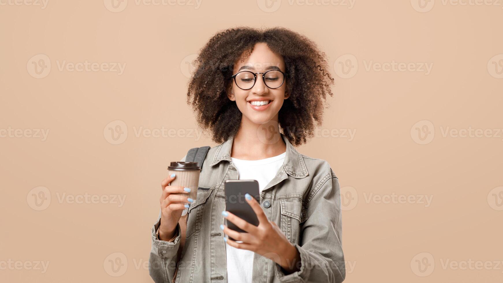 Modern zoomer blogger and social media. Happy excited african american curly woman in glasses with backpack, holds takeaway coffee and reads message in smartphone, isolated on light background photo