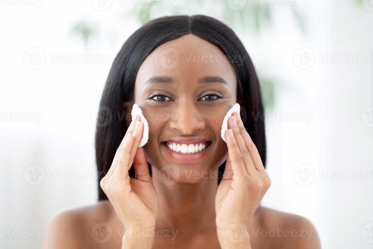 Smiling young african american woman doing daily morning skincare procedures, cleans face skin with natural lotion tonic using cotton disc sponge pad or remove makeup by micellar water at evening photo
