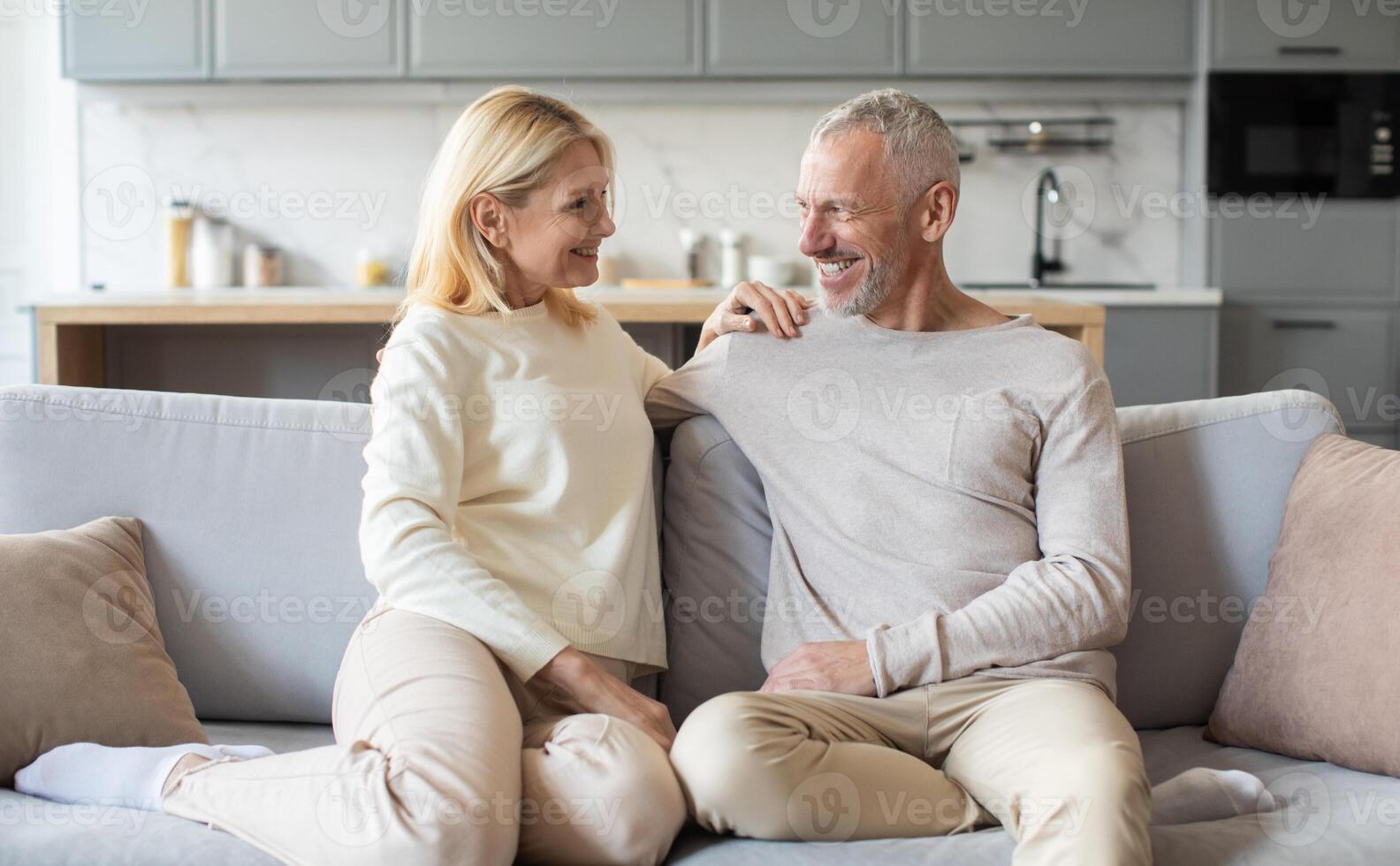A joyful couple relaxes on a comfortable sofa in a modern living room, sharing smiles and affection while dressed casually in light-colored clothing. The warm atmosphere fosters their connection. photo
