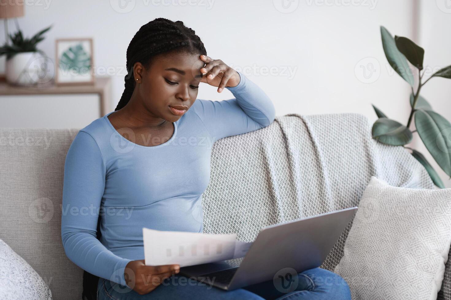 Remote Business. Pensive african woman checking papers and working with laptop at home, thoughtful black female freelancer busy with financial reports and documentation, sitting on sofa in living room photo