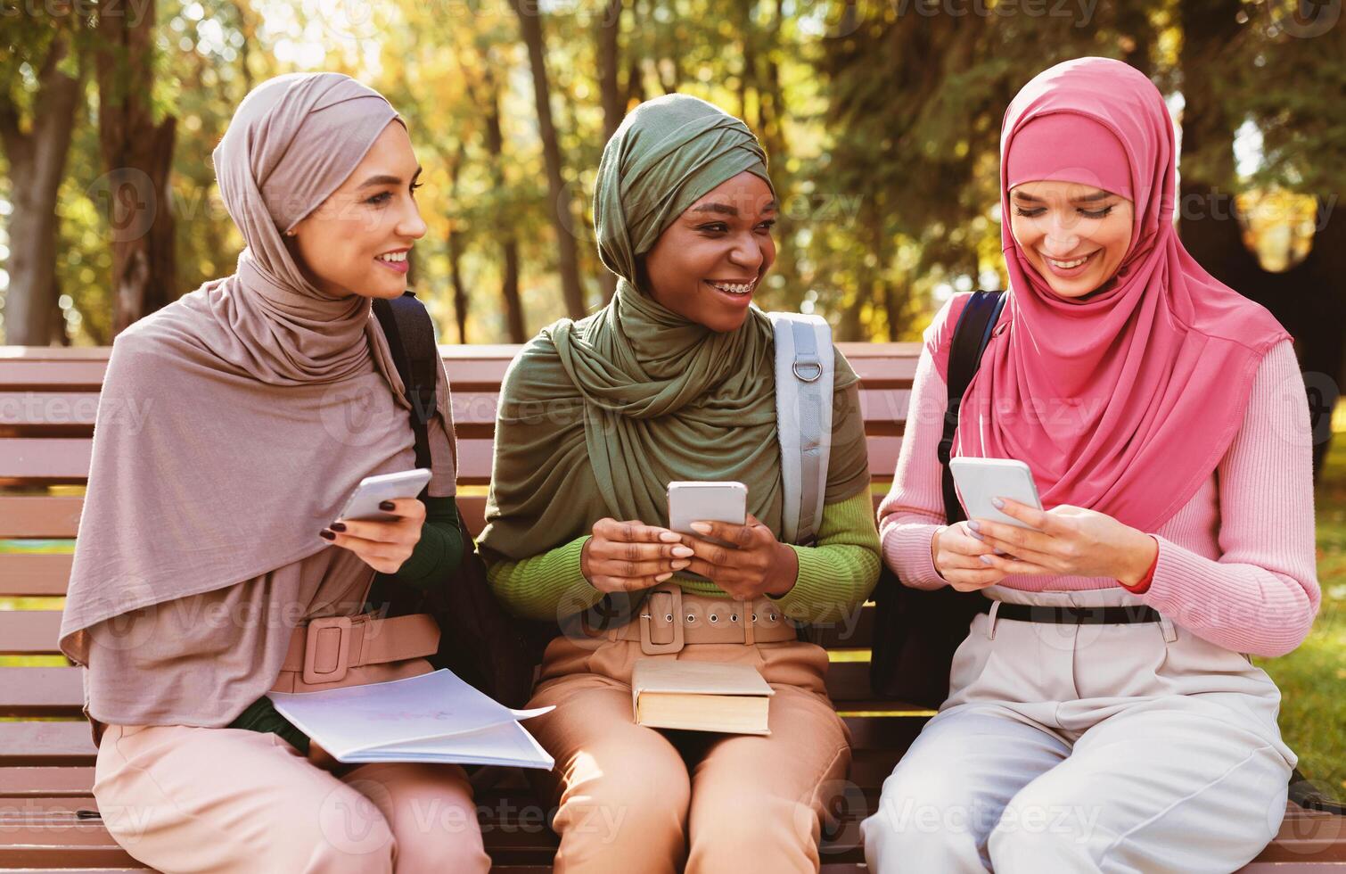 Three Modern Muslim Students Women Using Smartphones Sitting On Bench In Park Outdoors. Arabic Ladies In Hijab Texting On Phone Networking In Social Media. Educational Application, Gadget Lifestyle photo