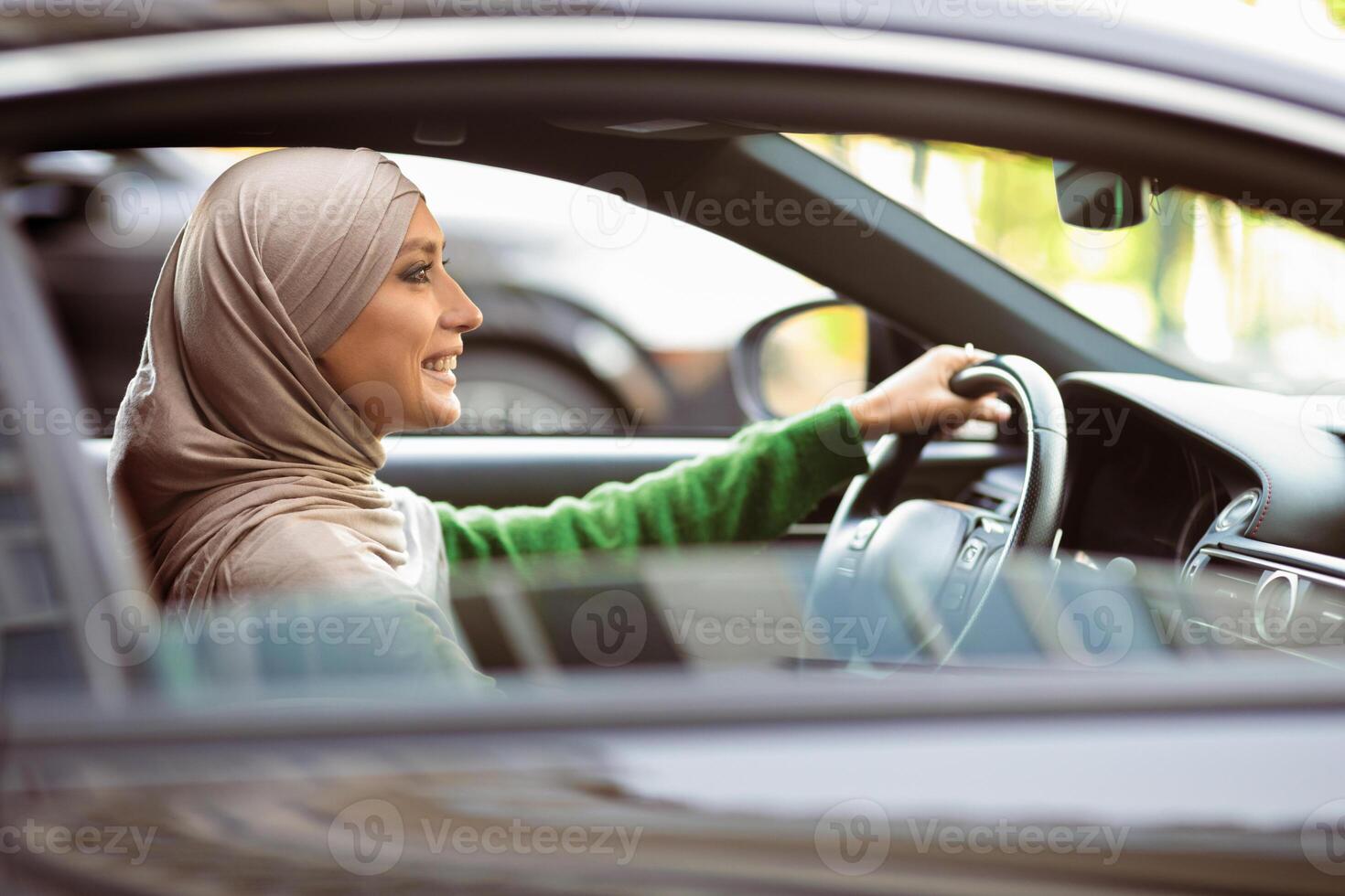 Female Driver. Side view profile portrait of positive islamic lady wearing headscarf driving her new luxury car. Smiling muslim woman in hijab holding steering wheel, view from outside photo