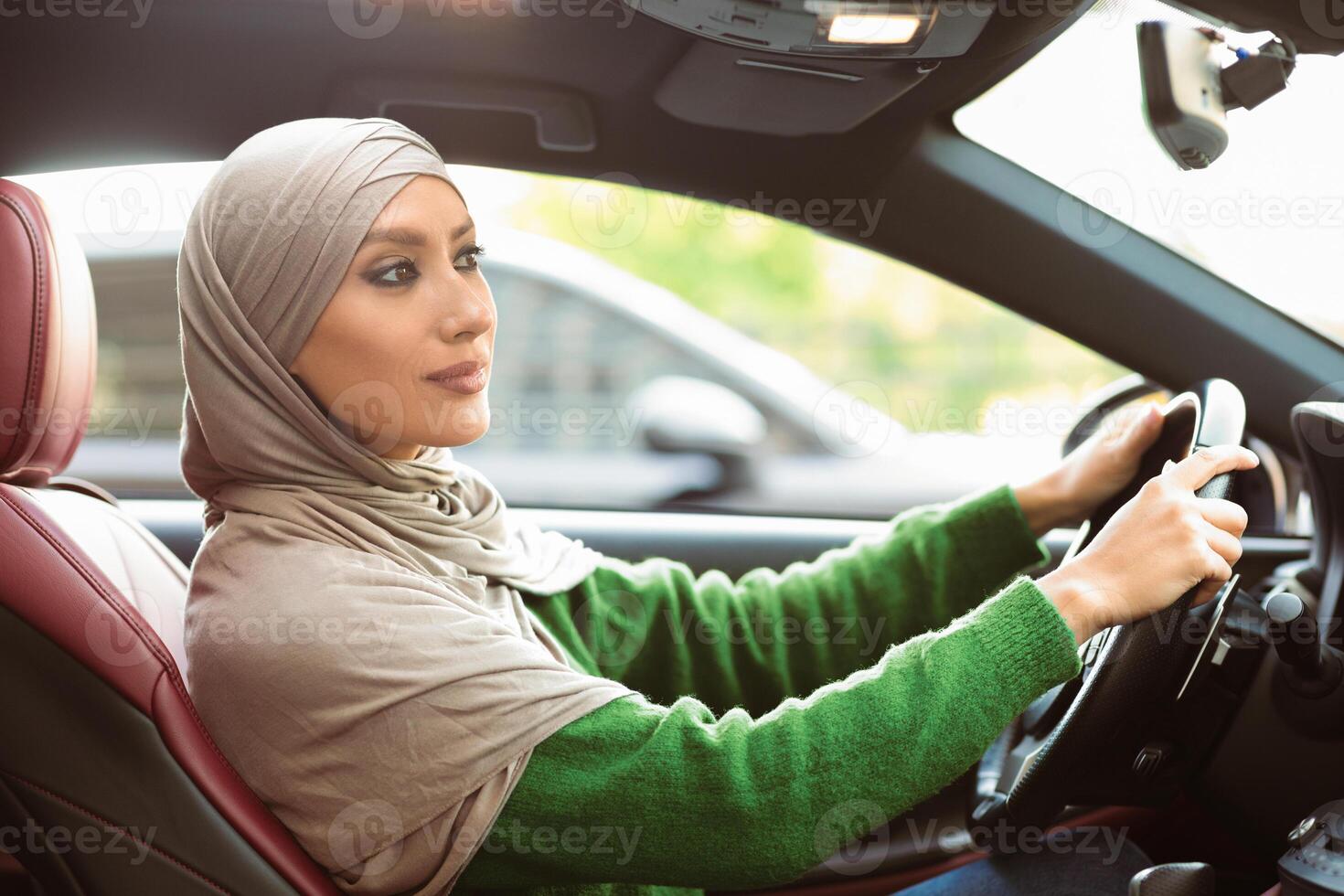 Female Driver. Side view portrait of confident islamic lady wearing headscarf driving her new auto. Young muslim woman in hijab holding steering wheel, view from the inside of luxury vehicle photo
